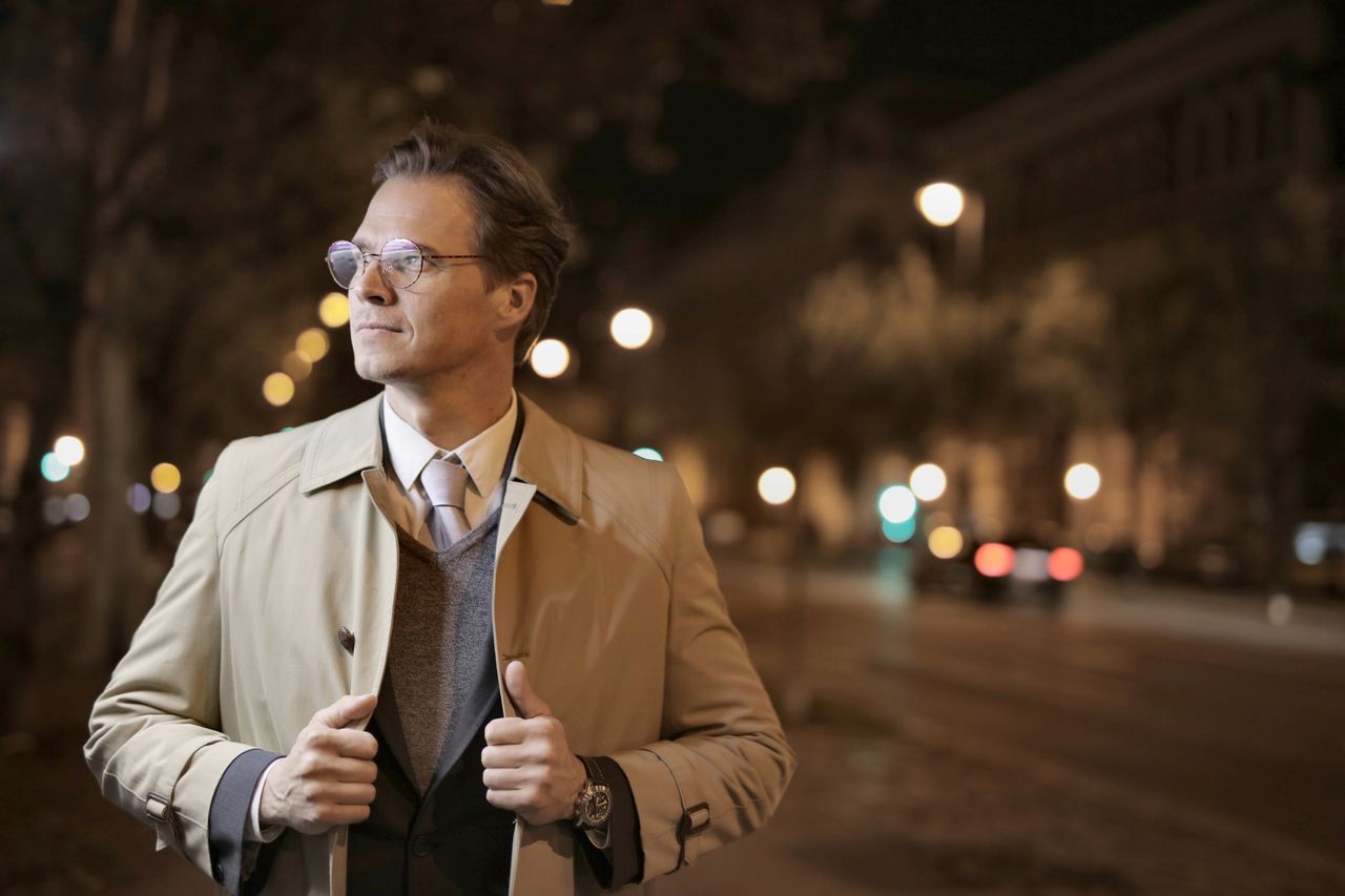 Confident adult male in formal wear standing on night street and holding collars of trench while looking away
